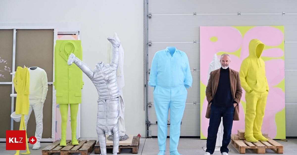 Ein Mann in einem braunen Mantel steht neben zwei großen Mannequins, die farbenfrohe Kleidung tragen. Ein Mannequin ist silber mit einem erhobenen Arm und das andere ist blau. Dahinter hängt ein großes rosa und gelbes Gemälde.