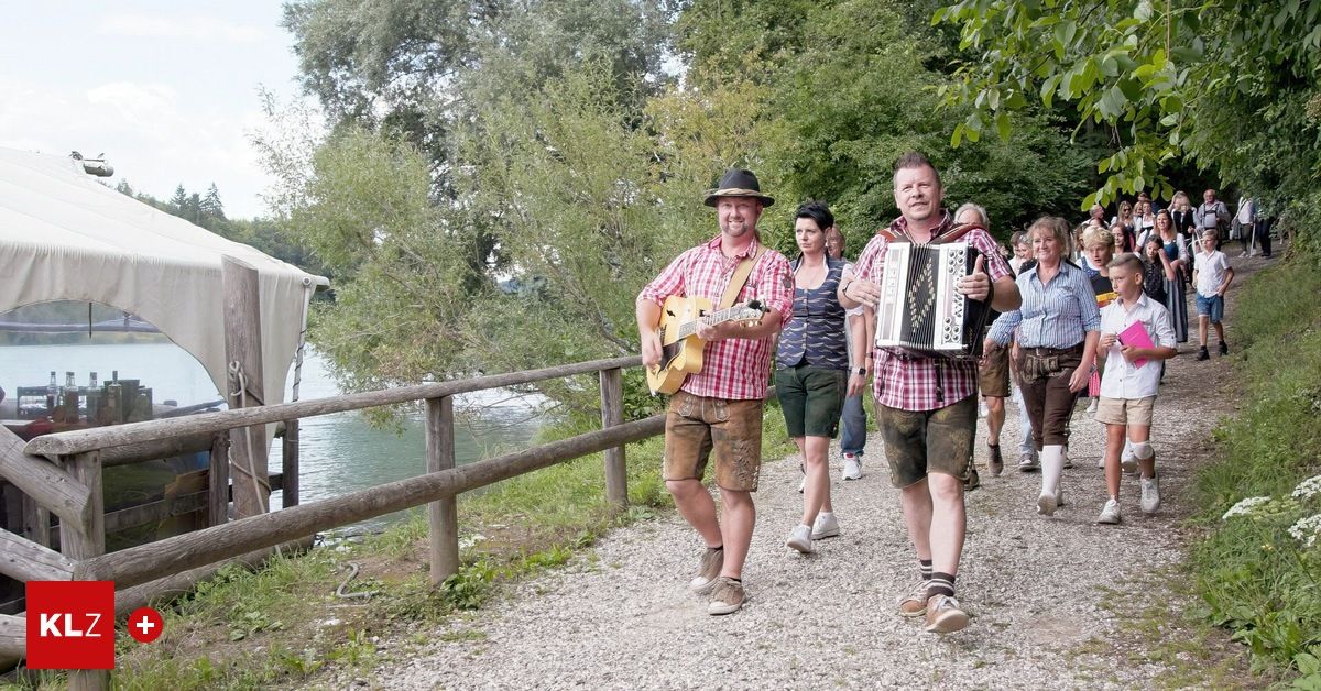 Eine Gruppe von Männern und Frauen in traditioneller Kleidung, die einen Schottenweg entlang neben einem Fluss geht. Ein Mann spielt Gitarre und ein anderer spielt Akkordeon.