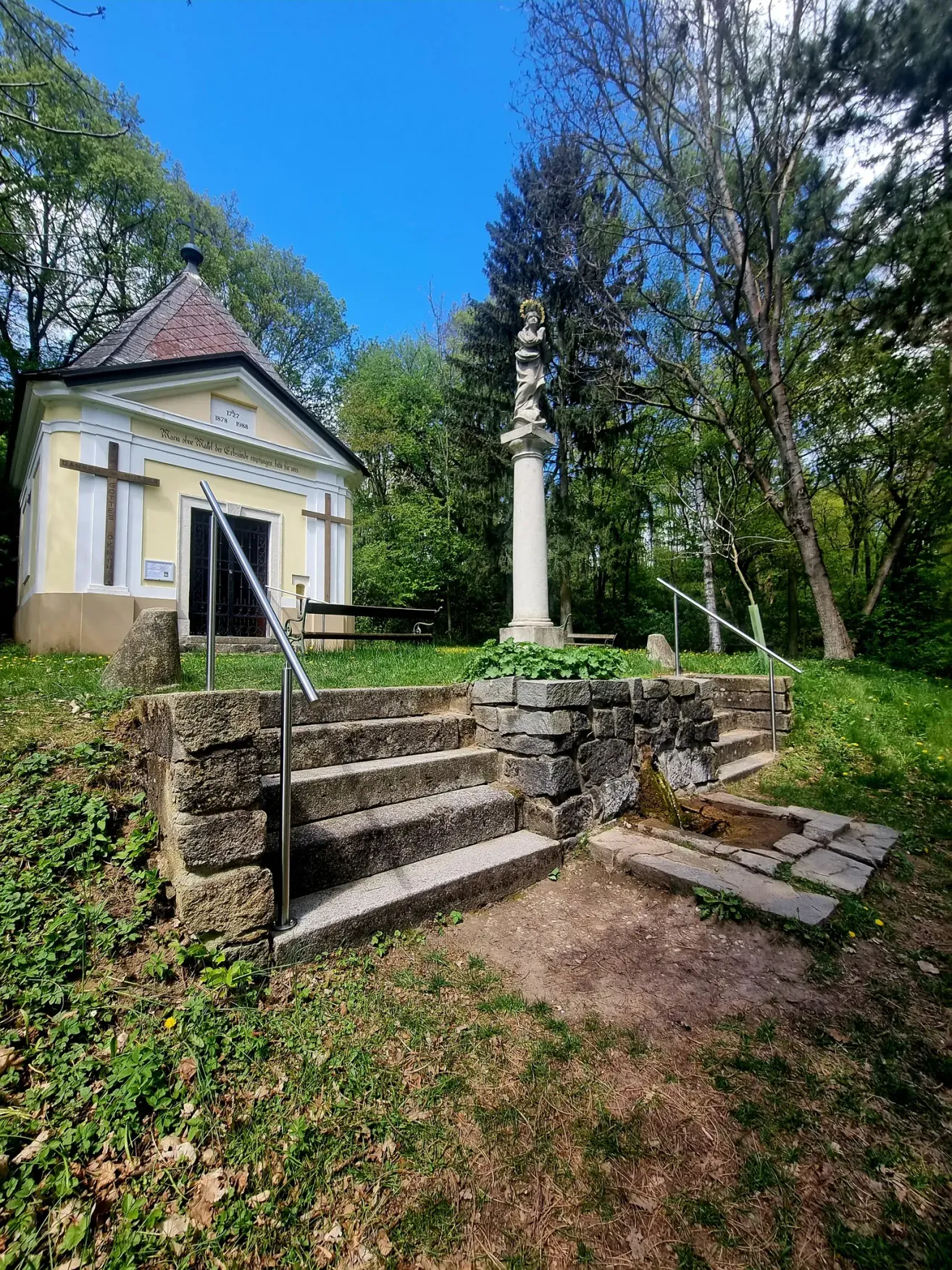 Eine kleine Kapelle mit einer Statue, Stufen und Handläufen, umgeben von Grün und einer Bank.