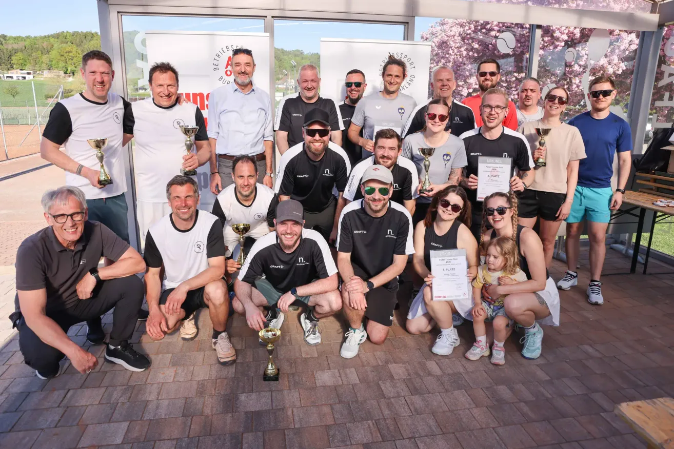Eine Gruppe von Männern und Frauen posiert mit ihren Trophäen auf einem Ziegelboden. Sie tragen alle Sonnenbrillen, Hüte und Sneakers. Einer von ihnen hält ein Zertifikat. Sie stehen vor einer Glastür.