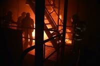 Firefighters stand near a burning building with a staircase leading to the upper floor. The fire illuminates the scene, casting shadows on the surrounding area.