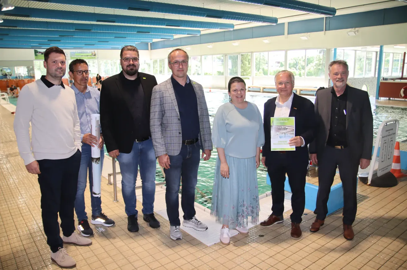 Eine Gruppe von Personen steht in einem Poolbereich, lächelt und hält ein Buch. Die Frau in der Mitte trägt ein Kleid und hat einen Ring am Finger. Der Mann auf der rechten Seite hält ein Buch und trägt eine Brille. Sie stehen vor einem Pool mit blauem Wasser.