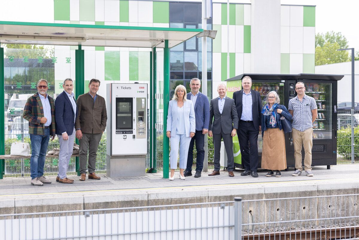 Several people in business attire stand on a platform in front of a ticket machine, with a building and vending machine in the background.