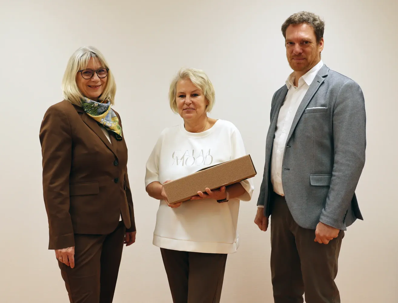 Drei Menschen stehen dicht beieinander vor einer weißen Wand und lächeln für ein Foto. Eine Frau hält eine braune Box und zwei Personen tragen formelle Kleidung.