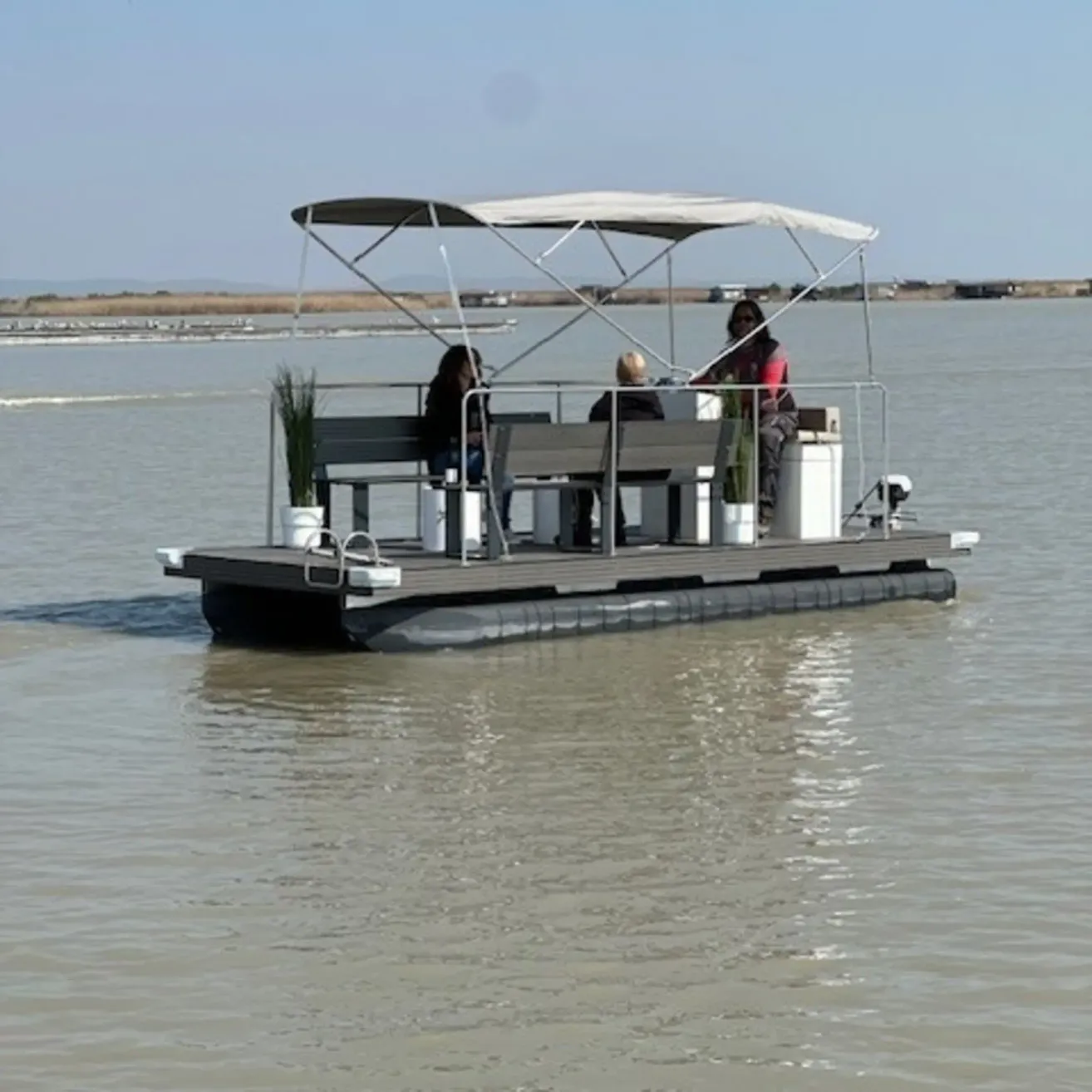 Three people sit on a pontoon boat with a canopy in a calm lake. There are plants in white pots on the deck.