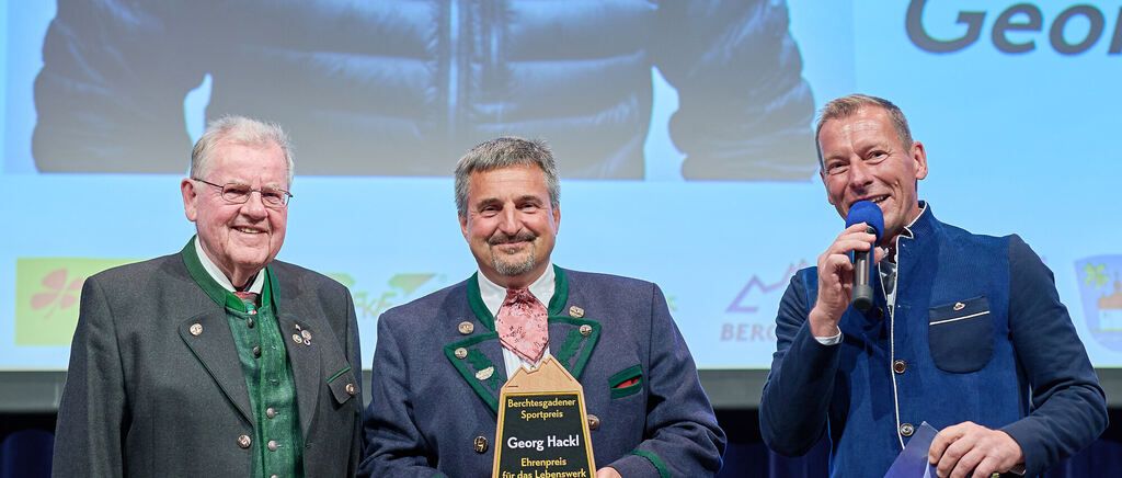 Three men stand together, smiling for a photo, with one holding a microphone and another holding a plaque. Behind them, a large screen displays the word 'Berg'.