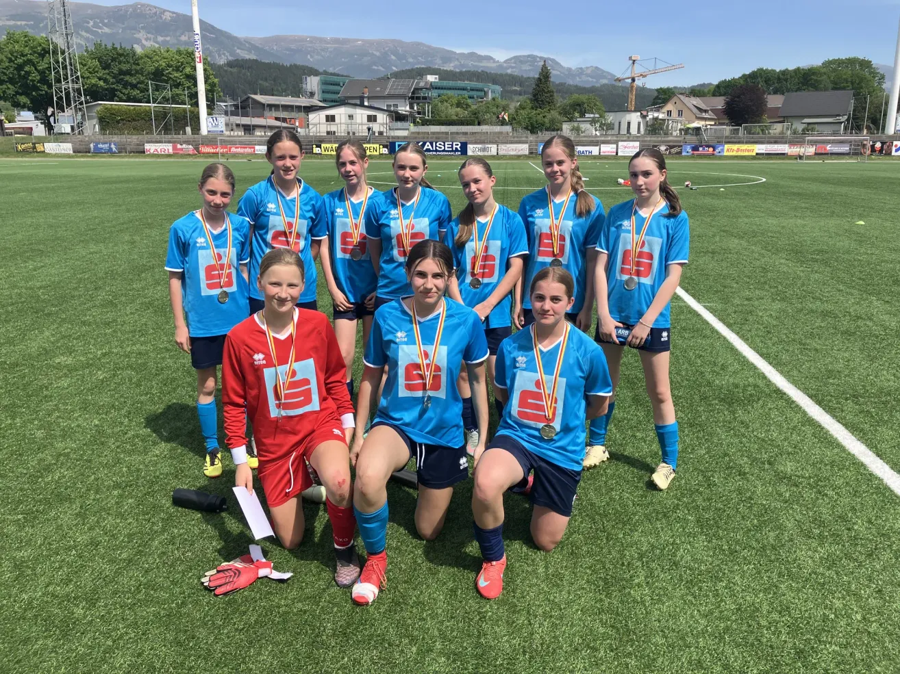 A group of young girls wearing blue uniforms pose for a picture on a soccer field. They have medals around their necks and some are kneeling.