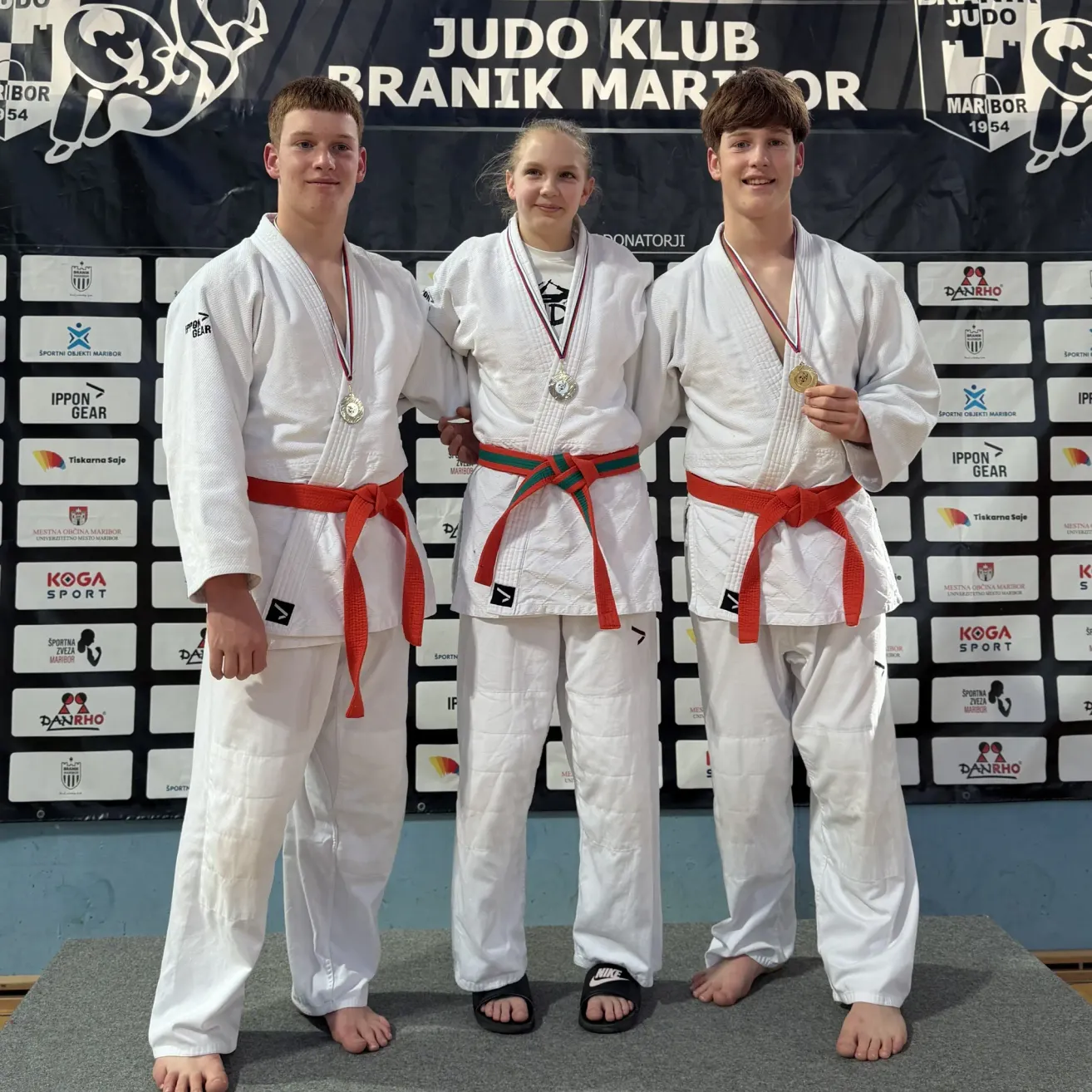Three young judokas stand on a stage wearing white uniforms and medals. They are posing for a photo with a backdrop that reads Judo Klub Branik Maribor.