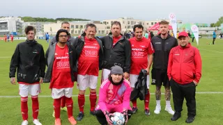 Eine Gruppe von Männern und Frauen posiert für ein Foto auf einem Fußballfeld, in roten und weißen Trikots, mit einem Fußball im Vordergrund.