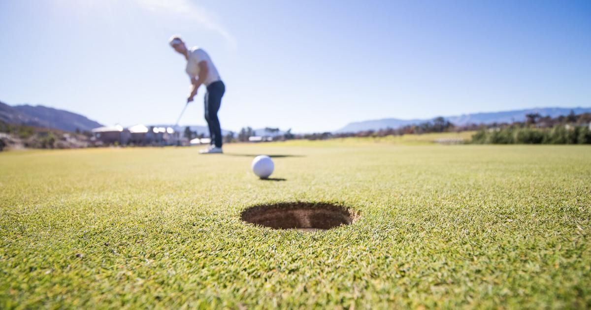 Ein Golfer steht auf einem Golfplatz und ist dabei, einen weißen Golfball in das Loch zu schlagen. Der Himmel ist klar.