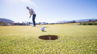 Ein Golfer steht auf einem Golfplatz und ist dabei, einen weißen Golfball in das Loch zu schlagen. Der Himmel ist klar.