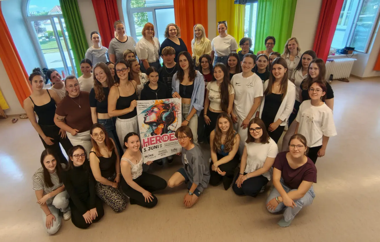 Eine Gruppe junger Menschen posiert für ein Foto in einem Raum mit einem Poster, das 'HEROE 3. JUNI' steht. Es gibt viele Menschen im Raum, alle lächeln für die Kamera. Der Raum ist mit farbenfrohen Vorhängen dekoriert.