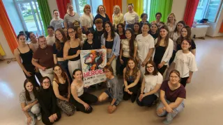 Eine Gruppe junger Menschen posiert für ein Foto in einem Raum mit einem Poster, das 'HEROE 3. JUNI' steht. Es gibt viele Menschen im Raum, alle lächeln für die Kamera. Der Raum ist mit farbenfrohen Vorhängen dekoriert.