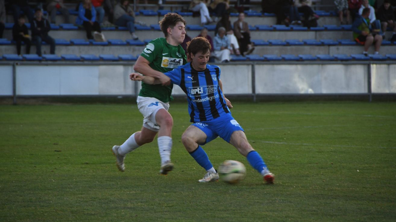 Zwei Fußballspieler spielen auf einem Feld, einer trägt Grün und der andere Blau. Der blaue Spieler tritt den Ball, während der Grüne versucht, ihn zu blockieren.