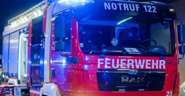 Ein rotes Notfallfahrzeug mit blauen Lichtern steht in einem dunklen Bereich. Es hat das Wort 'Feuerwehr' und die Nummer '12' auf der Vorderseite. Die Lichter des Fahrzeugs sind eingeschaltet.