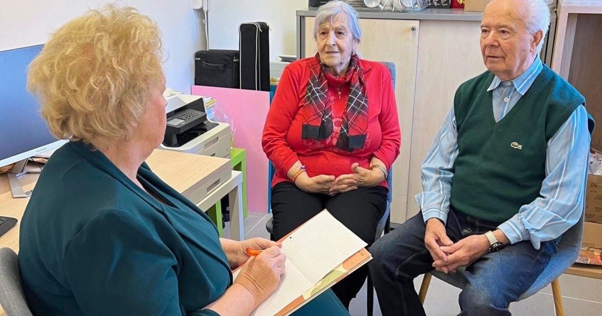 Drei Personen befinden sich in einem Innenraum, wahrscheinlich einem Büro. Eine Frau mit einem Notizbuch macht Notizen. Eine ältere Frau sitzt mit den Händen auf dem Bauch. Ein anderer Mann sitzt neben ihr.