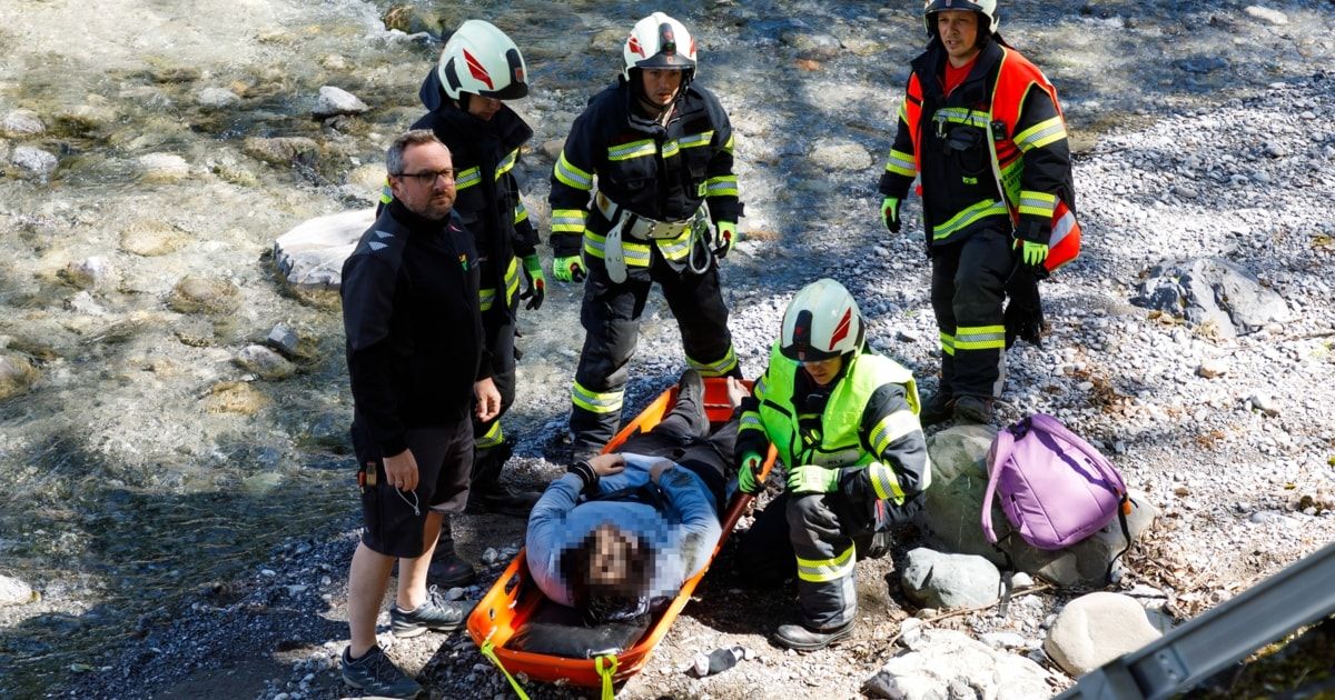 A rescue team is carrying a person on a stretcher, with one person blurred. A river flows nearby, and rocks are scattered around.