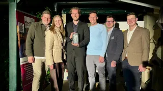 Fünf Personen, drei Männer und zwei Frauen, posieren für ein Foto und halten eine Trophäe. Sie lächeln und scheinen eine Leistung zu feiern.