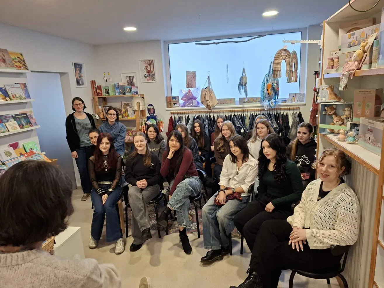 Eine Gruppe von Frauen sitzt in einem Kreis in einem Raum, wahrscheinlich um ein Thema zu diskutieren. Sie sind von Regalen mit Büchern und ausgestellten Gegenständen umgeben. Ein Fenster zeigt weitere Gegenstände und einen Kleiderständer.