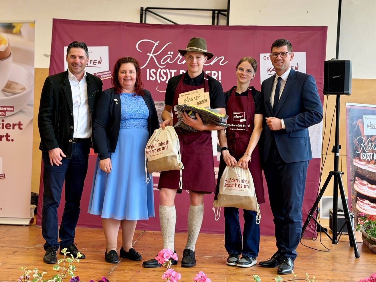 Fünf Personen stehen auf einer Bühne. Die zentrale Figur hält ein Paket. Der Hintergrund zeigt 'Karnten IS(S)T BERI'. Der Mann auf der rechten Seite trägt eine Krawatte. Die beiden Frauen halten Taschen.