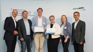 Fünf Menschen stehen lächelnd vor einer weißen Wand. Sie sind in formeller Kleidung gekleidet. Die beiden Frauen halten Auszeichnungen und der Mann auf der linken Seite ein Zertifikat. Hinter ihnen befindet sich eine Wand mit Text und Schatten.