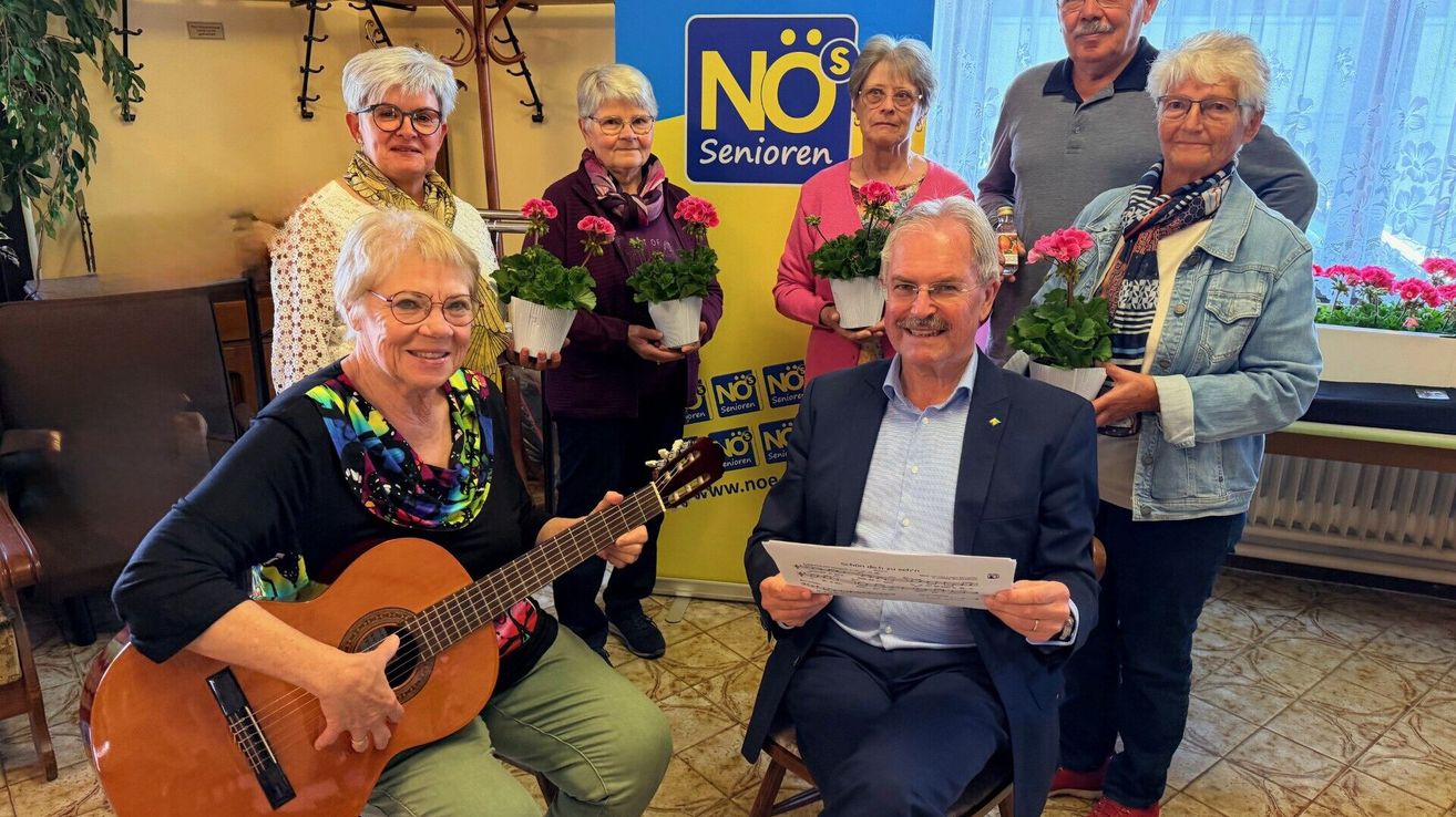 Gruppe älterer Menschen, die für ein Foto posieren, einige mit Gitarren und Blumen. Ein Mann liest ein Blatt Papier neben einem Schild mit 'NO's Senioren'.