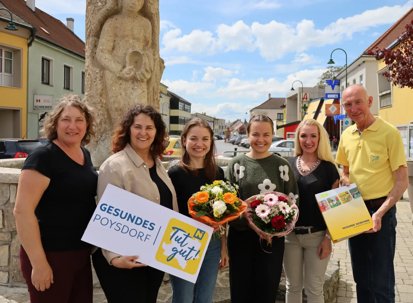 Fünf Frauen und ein Mann stehen vor einer Statue, halten Blumensträuße und Schilder. Die Frauen lächeln. Dahinter ist eine Straße mit geparkten Autos und Gebäuden.