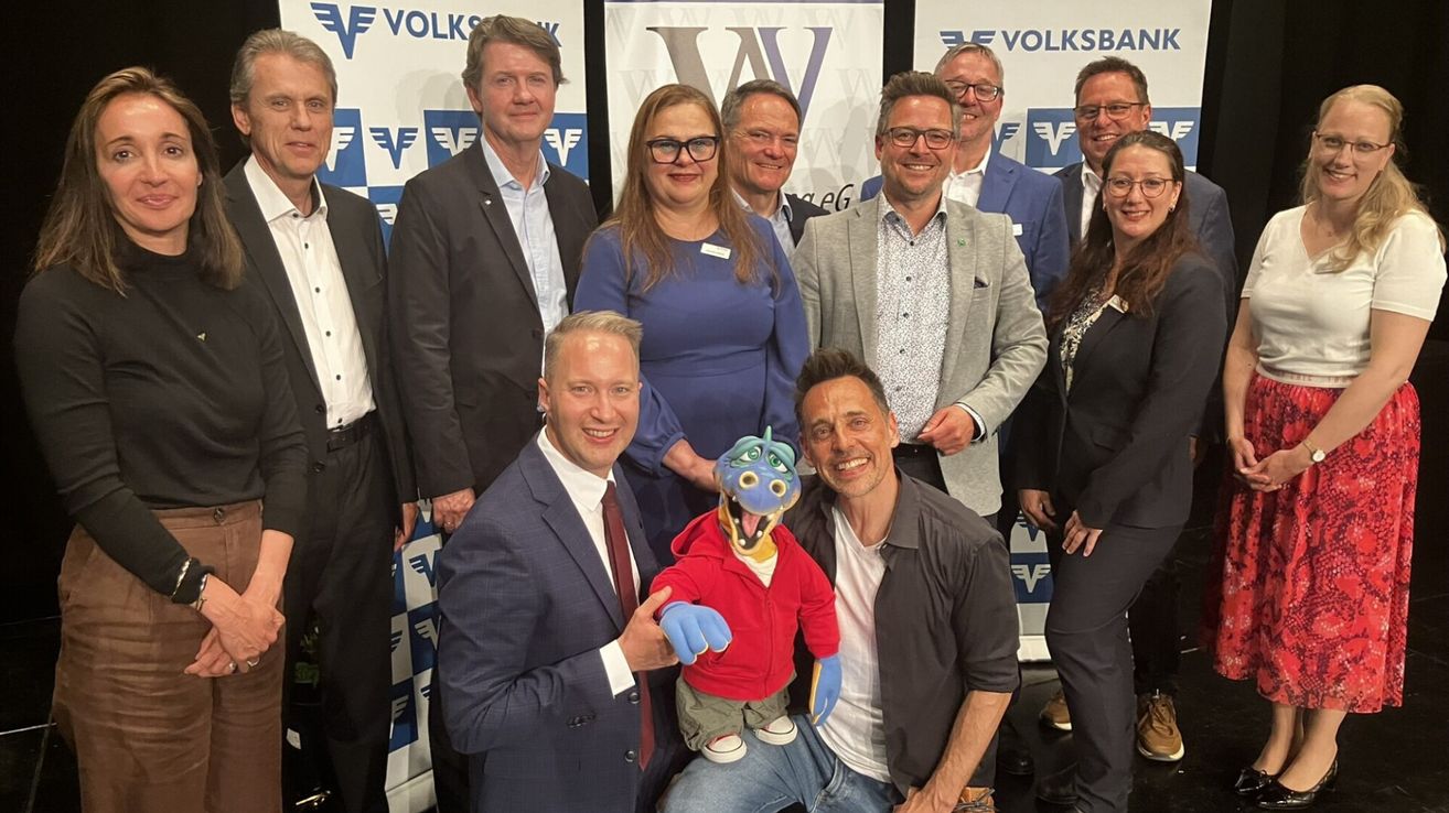 Eine Gruppe von Menschen posiert für ein Foto, lächelt und steht hinter einem Banner. Ein Mann hält eine Puppe.