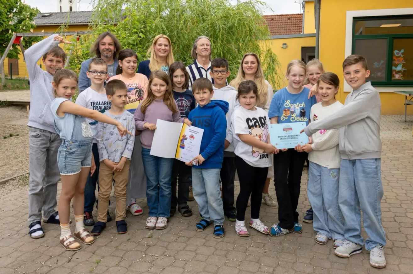 A group of children and adults stand outside, some holding certificates. They are dressed casually, standing in front of a house with yellow walls and a roof. Trees and plants are in the background.