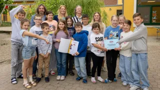 Eine Gruppe von Kindern und Erwachsenen steht draußen, einige halten Urkunden. Sie sind lässig gekleidet und stehen vor einem Haus mit gelben Wänden und einem Dach. Bäume und Pflanzen sind im Hintergrund.