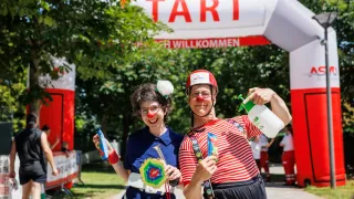 Zwei Clowns, einer mit einer Trompete, stehen unter einem Banner mit der Aufschrift 'TART' und halten Sprühflaschen. Bäume und andere Leute sind im Hintergrund.