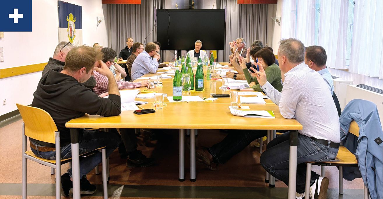 A group of people in a meeting room with a long table, bottles, papers, and a television in the background.