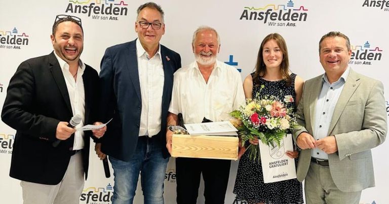 Fünf Personen stehen lächelnd nebeneinander, halten eine Box und einen Blumenstrauß. Der Mann in der Mitte hält eine Box mit einem Band und einer Karte.