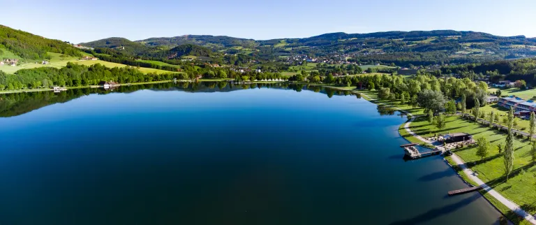 Ein ruhiger See mit Bergreflexion, umgeben von üppigem Grün und einer kleinen Stadt in der Ferne.