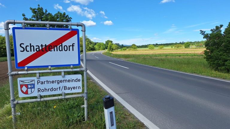 Ein Straßenschild für Endorrf, ein Dorf in Bayern, steht am Straßenrand, mit einem weiten grünen Feld, das sich unter einem klaren blauen Himmel erstreckt.