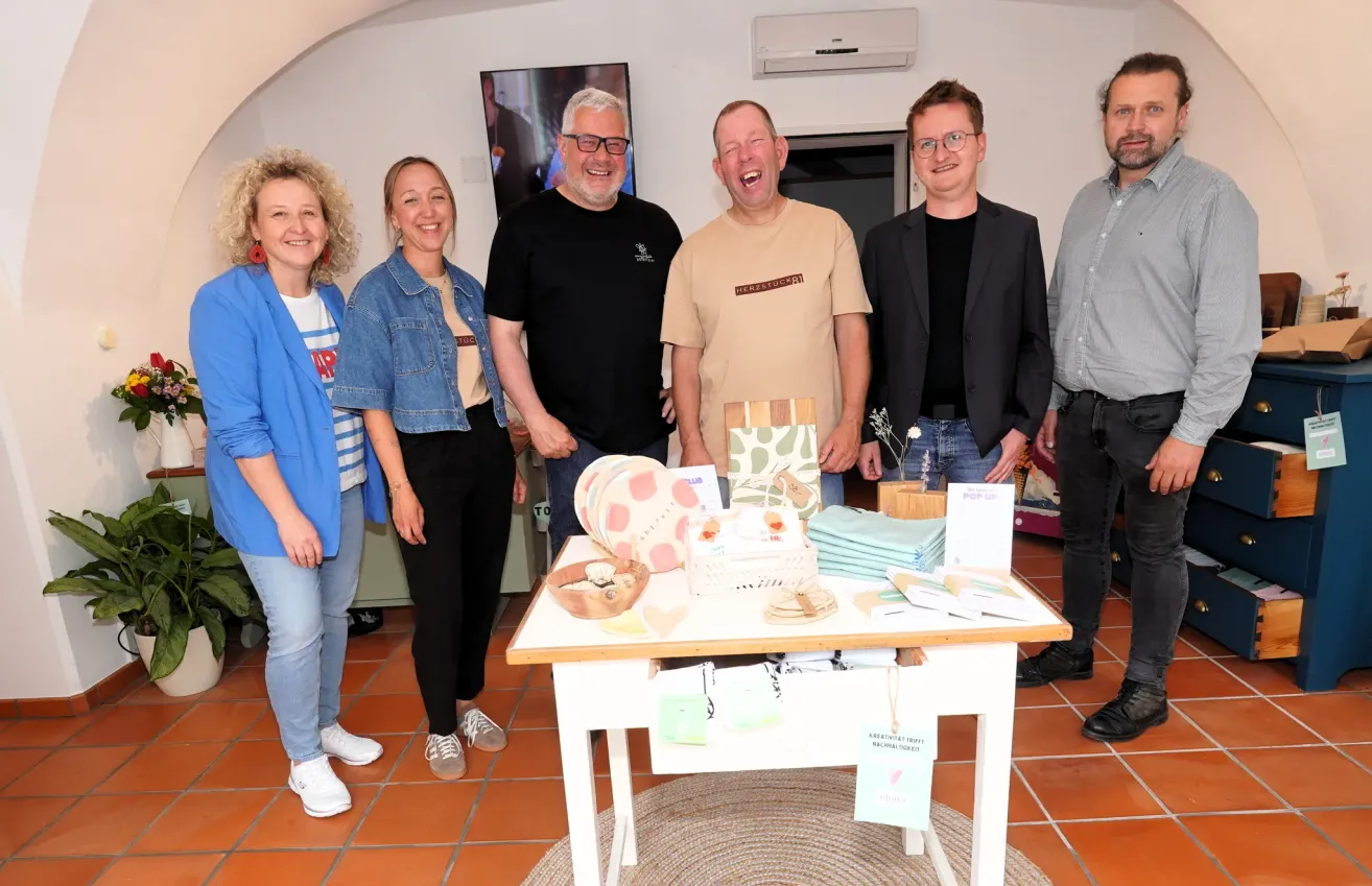 Fünf Personen stehen in einem Raum mit lächelnden Gesichtern. Ein Tisch davor hat Produkte darauf. Zwei Männer tragen Brillen. Ein Fernseher ist an der Wand montiert.