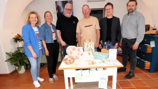 Fünf Personen stehen in einem Raum mit lächelnden Gesichtern. Ein Tisch davor hat Produkte darauf. Zwei Männer tragen Brillen. Ein Fernseher ist an der Wand montiert.