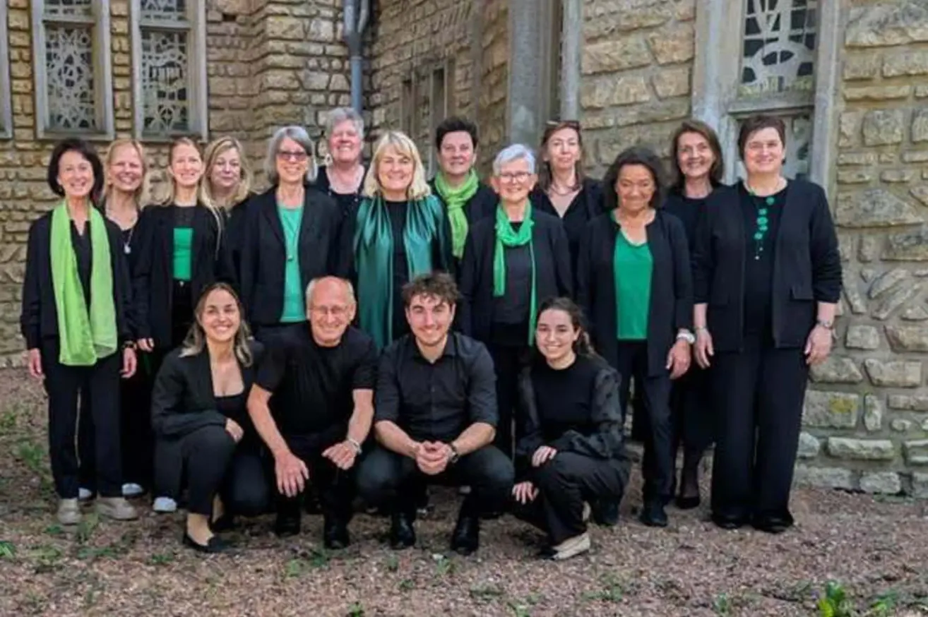 Ein Chor von Menschen in Grün und Schwarz steht vor einem Steingebäude. Einige tragen Brillen und Schals. Einige knien oder hockten sich hin.