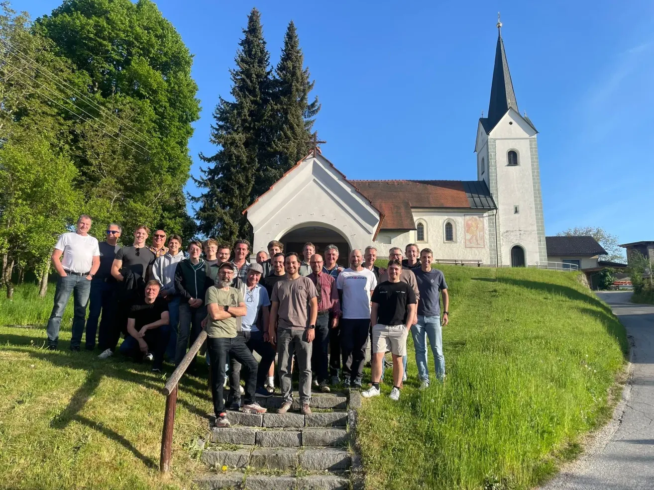 Eine Gruppe von Männern posiert für ein Foto vor einer Kirche mit einem Turm und Bäumen dahinter.