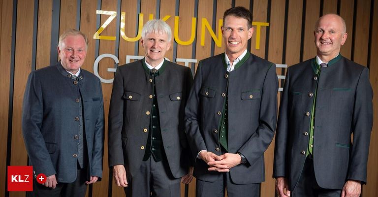 Vier Männer in traditioneller Kleidung stehen vor einer Holzwand mit gelben Buchstaben, die 'Zukunft' bilden. Sie lächeln und scheinen für ein Foto zu posieren.