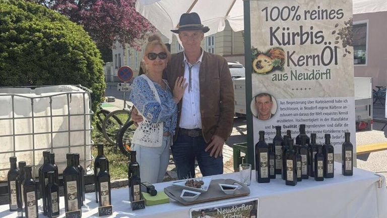 Ein Mann und eine Frau posieren an einem Tisch mit Ölflaschen und lächeln. Dahinter bewirbt ein Schild 100% Kürbis-Ke.