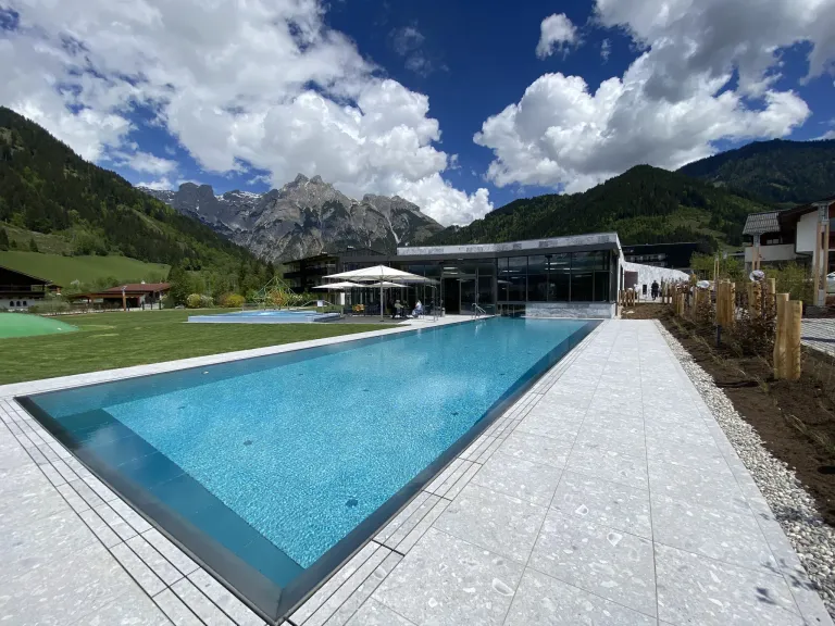 Ein großer Swimmingpool vor einem Gebäude mit Bergen im Hintergrund. Der Pool ist von Sonnenschirmen und einer grünen Wiese umgeben.