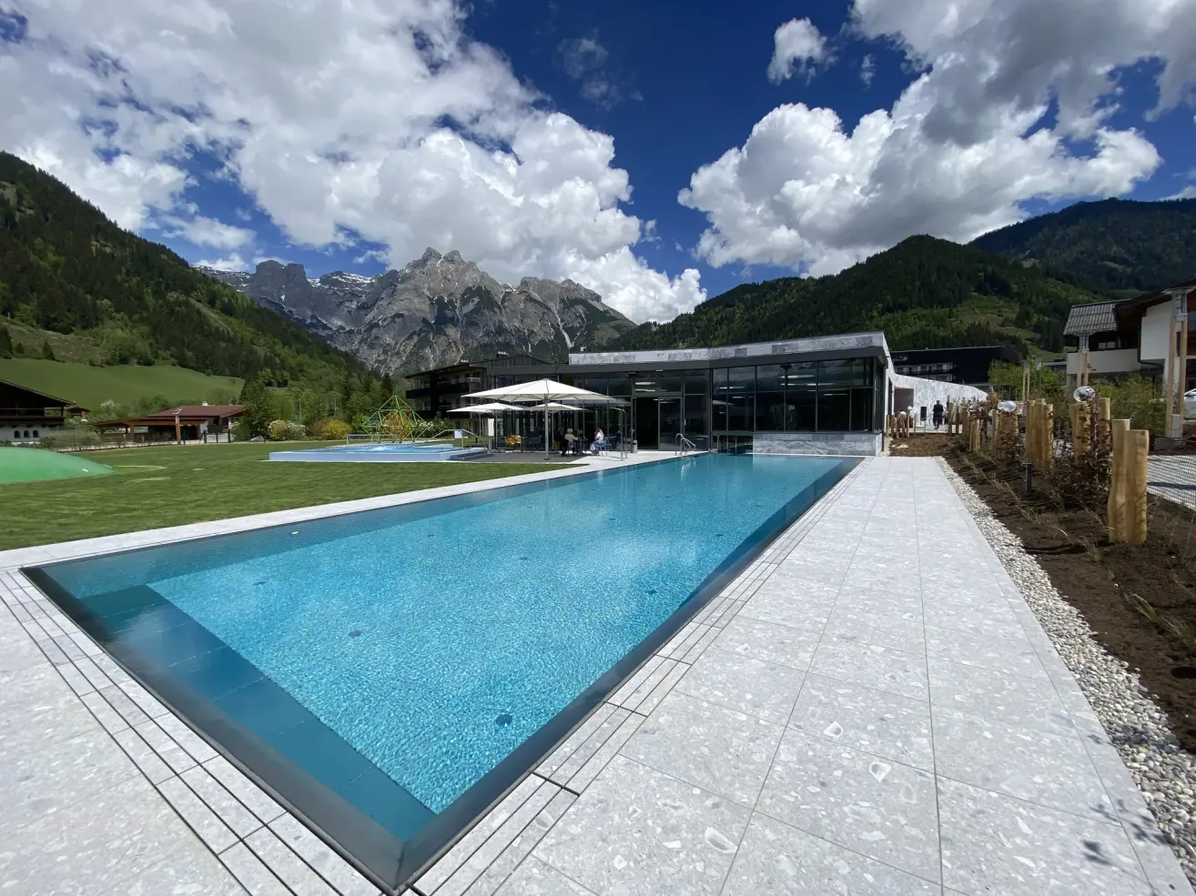 Ein großer Swimmingpool vor einem Gebäude mit Bergen im Hintergrund. Der Pool ist von Sonnenschirmen und einer grünen Wiese umgeben.