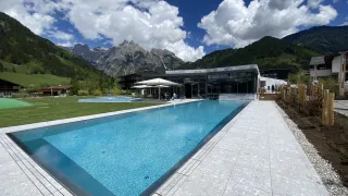 Ein großer Swimmingpool vor einem Gebäude mit Bergen im Hintergrund. Der Pool ist von Sonnenschirmen und einer grünen Wiese umgeben.