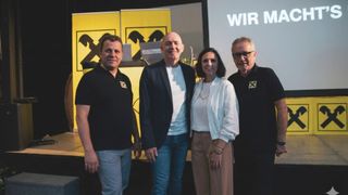 Vier Personen posieren für ein Foto vor einem gelben Hintergrund mit dem Wort Wir. Der Mann links trägt eine Brille.