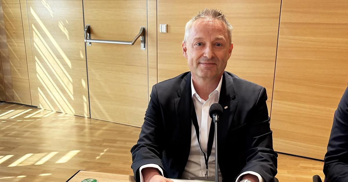 Ein Mann in Anzug sitzt an einem Schreibtisch mit Mikrofon, lächelt und schaut nach vorne, möglicherweise in einem Besprechungsraum oder Konferenzsaal.
