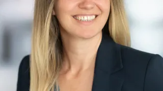 Eine Frau mit blonden Haaren und einem Nasenring lächelt in die Kamera. Sie trägt ein blaues Oberteil und einen schwarzen Blazer.