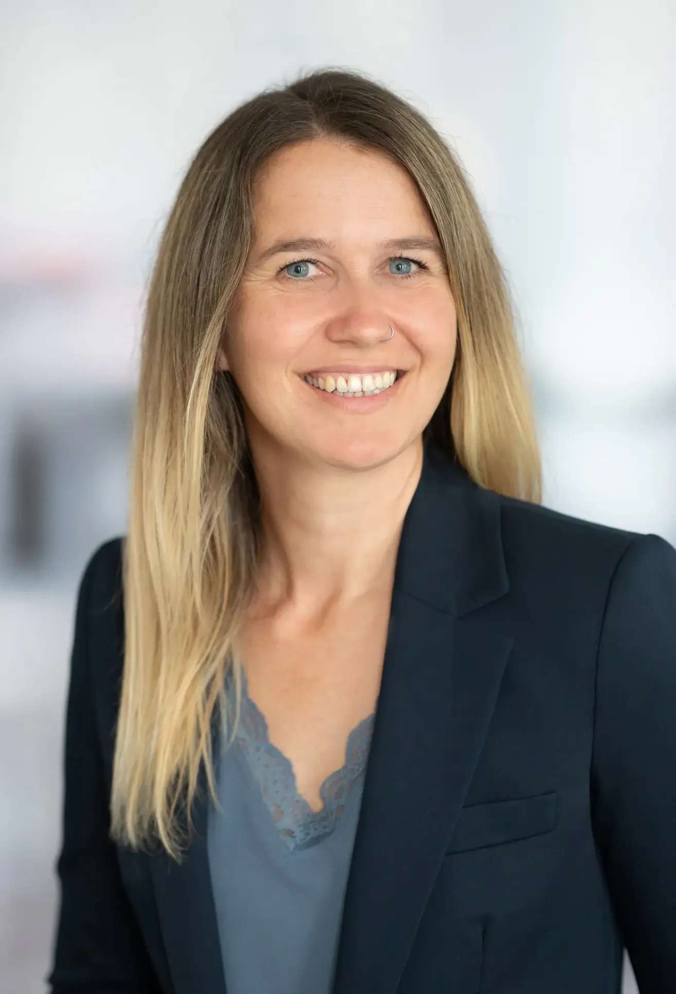 Eine Frau mit blonden Haaren und einem Nasenring lächelt in die Kamera. Sie trägt ein blaues Oberteil und einen schwarzen Blazer.