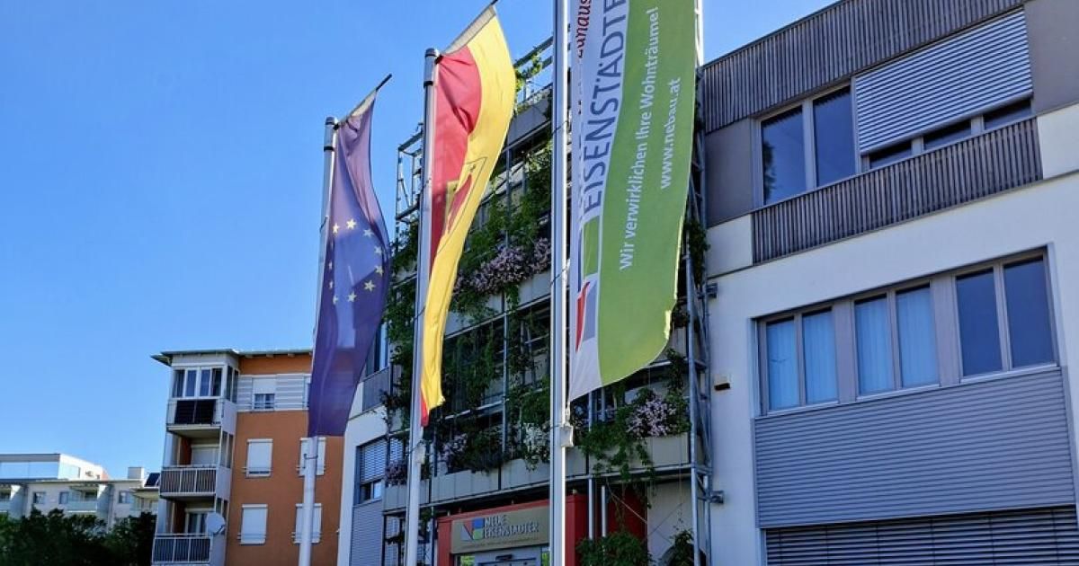 Drei Flaggen wehen vor einem modernen Gebäude mit Balkon. Eine grüne Flagge hat Text und eine Website. Eine gelb-rote Flagge steht neben ihr, und eine blaue Flagge ist links.