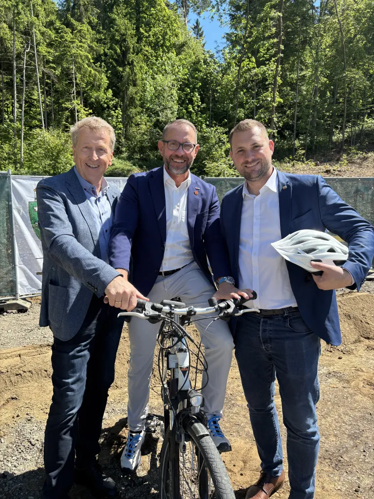 Three men in suits and glasses stand next to a bicycle. One man holds a helmet. They appear to be posing for a photograph at a construction site. Trees and a fence are in the background.